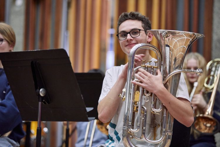 A boy in a white t-shirt holds a tuba and smiles at the camera